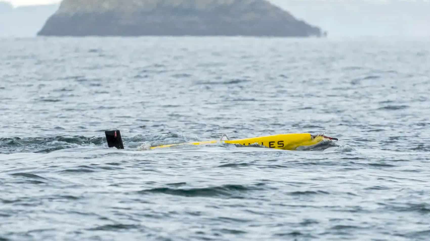 A yellow towed sonar array floating on open water near a rocky island.