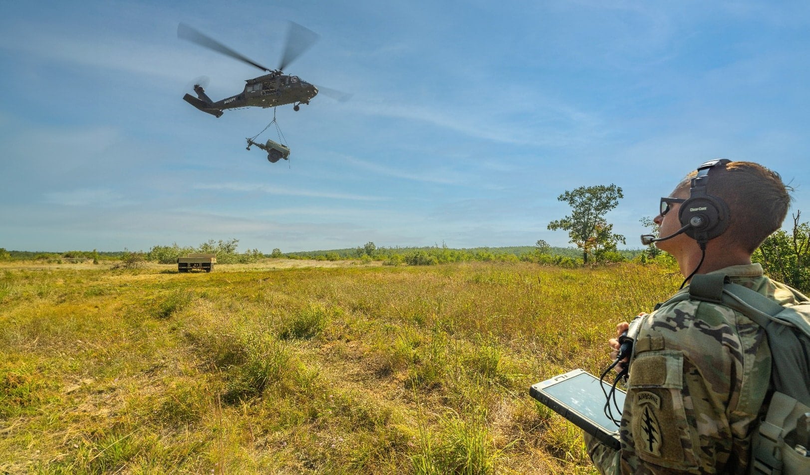 Soldier holds a tablet in a grass field as two helicopters pass overhead.