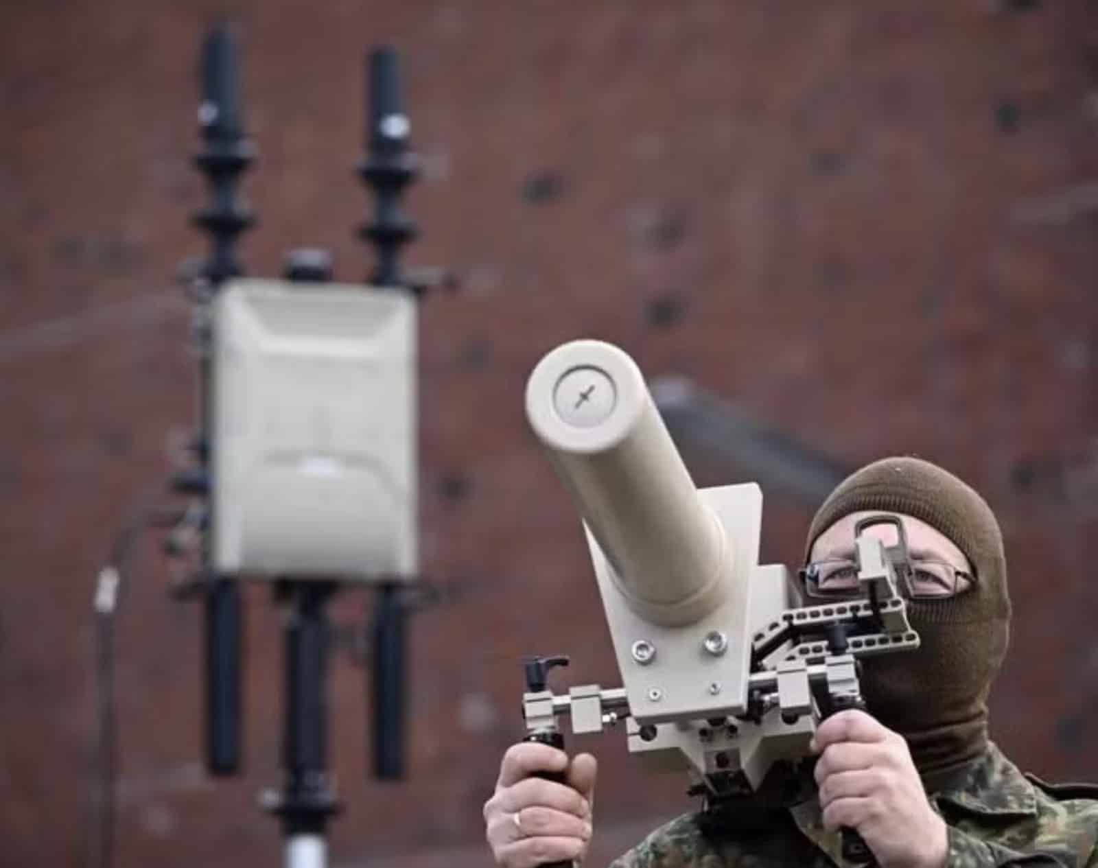 Masked soldier aims a shoulder-held counter-drone jammer beside a directional antenna mast.