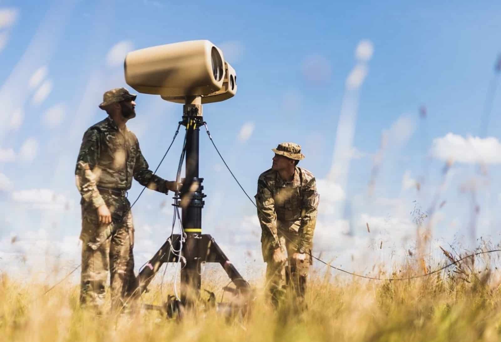 Two soldiers setting up a counter-drone sensor mast in a grassy field.
