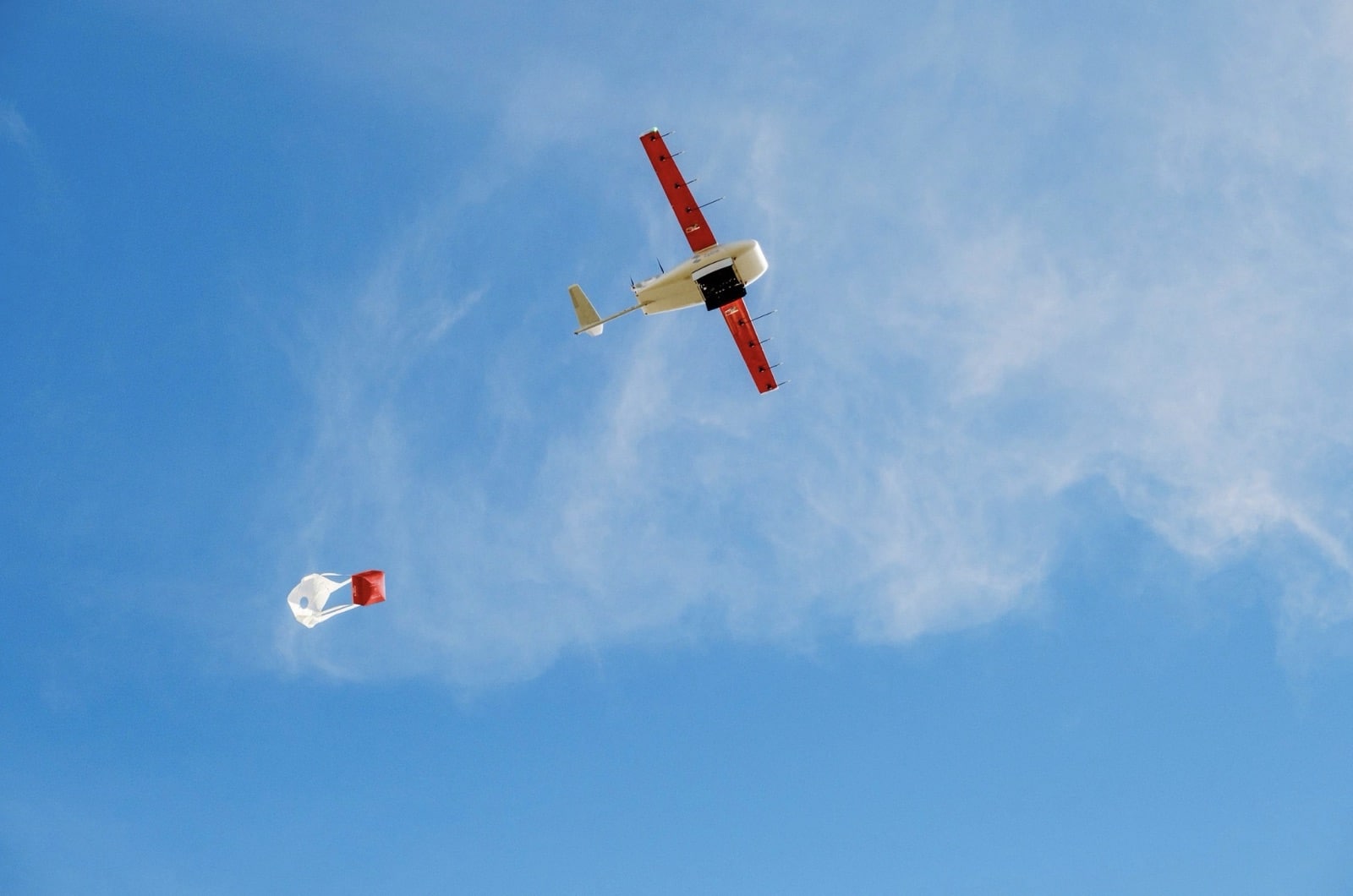 Zipline fixed-wing drone in blue sky lowering a package on a tether.