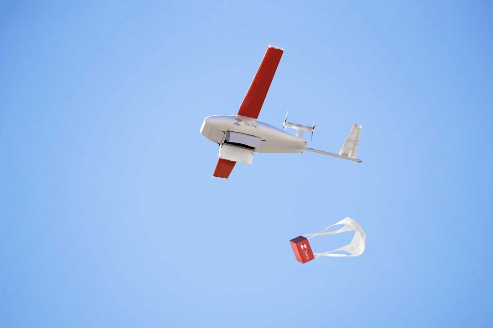 A delivery aircraft banks overhead as its package descends beneath a small parachute.