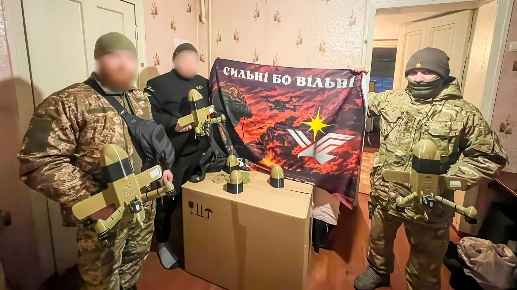 Soldiers with blurred faces stand beside supply boxes and a unit flag.