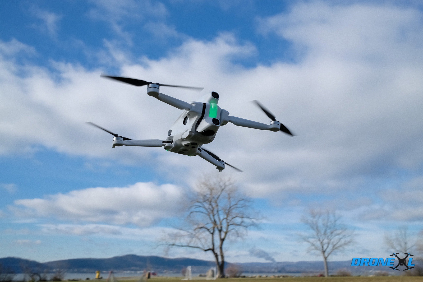 Folding camera drone hovering against a blue sky above bare winter trees.