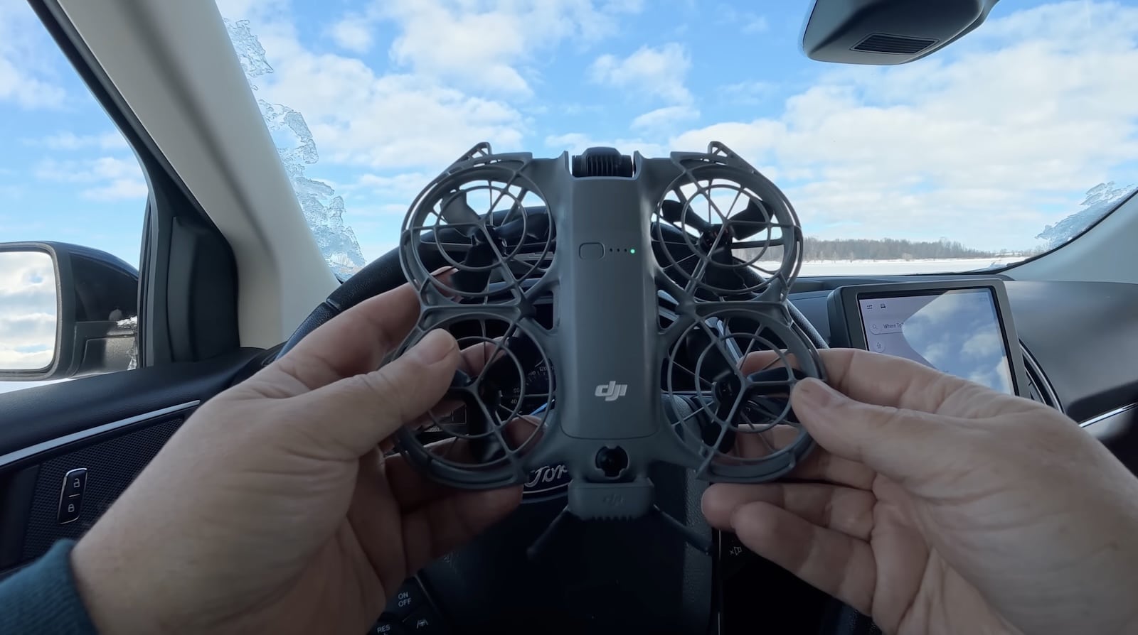 Hands hold a small ducted drone inside a car above a snowy landscape.