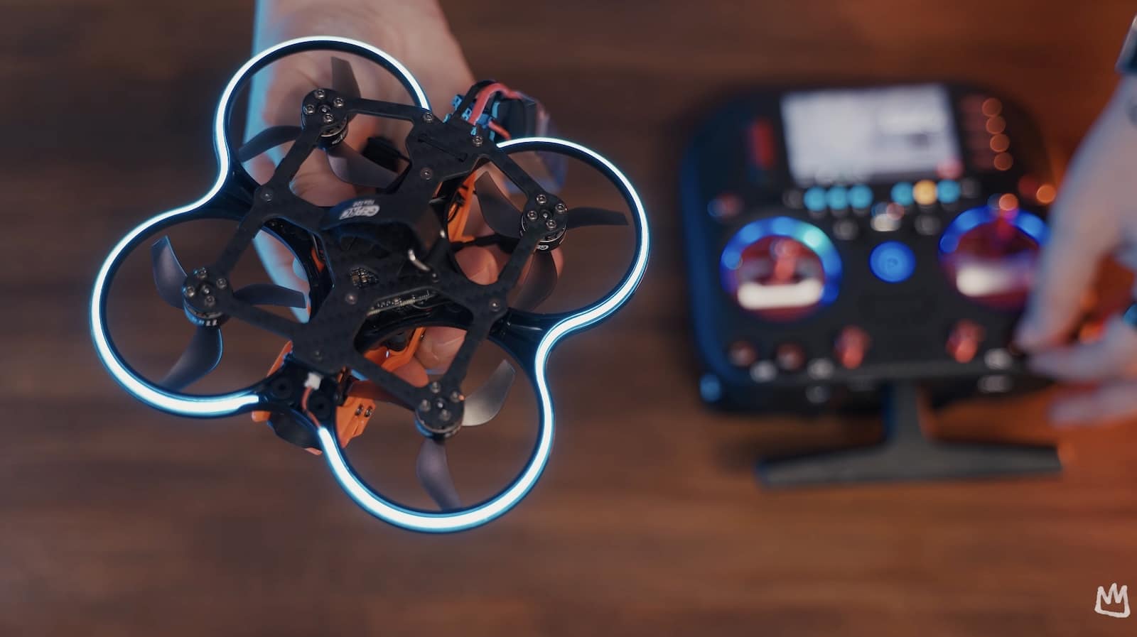 A hand holds the micro cinewhoop, LED-lit ducts glowing, beside its radio transmitter.