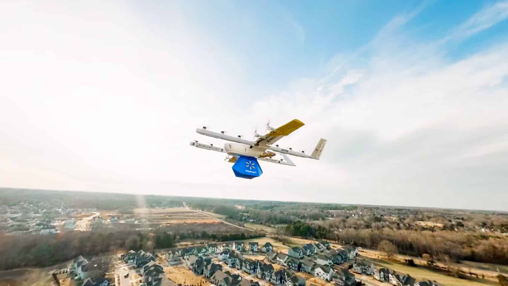 A Wing aircraft carries a blue Walmart package over a suburban neighborhood at altitude.