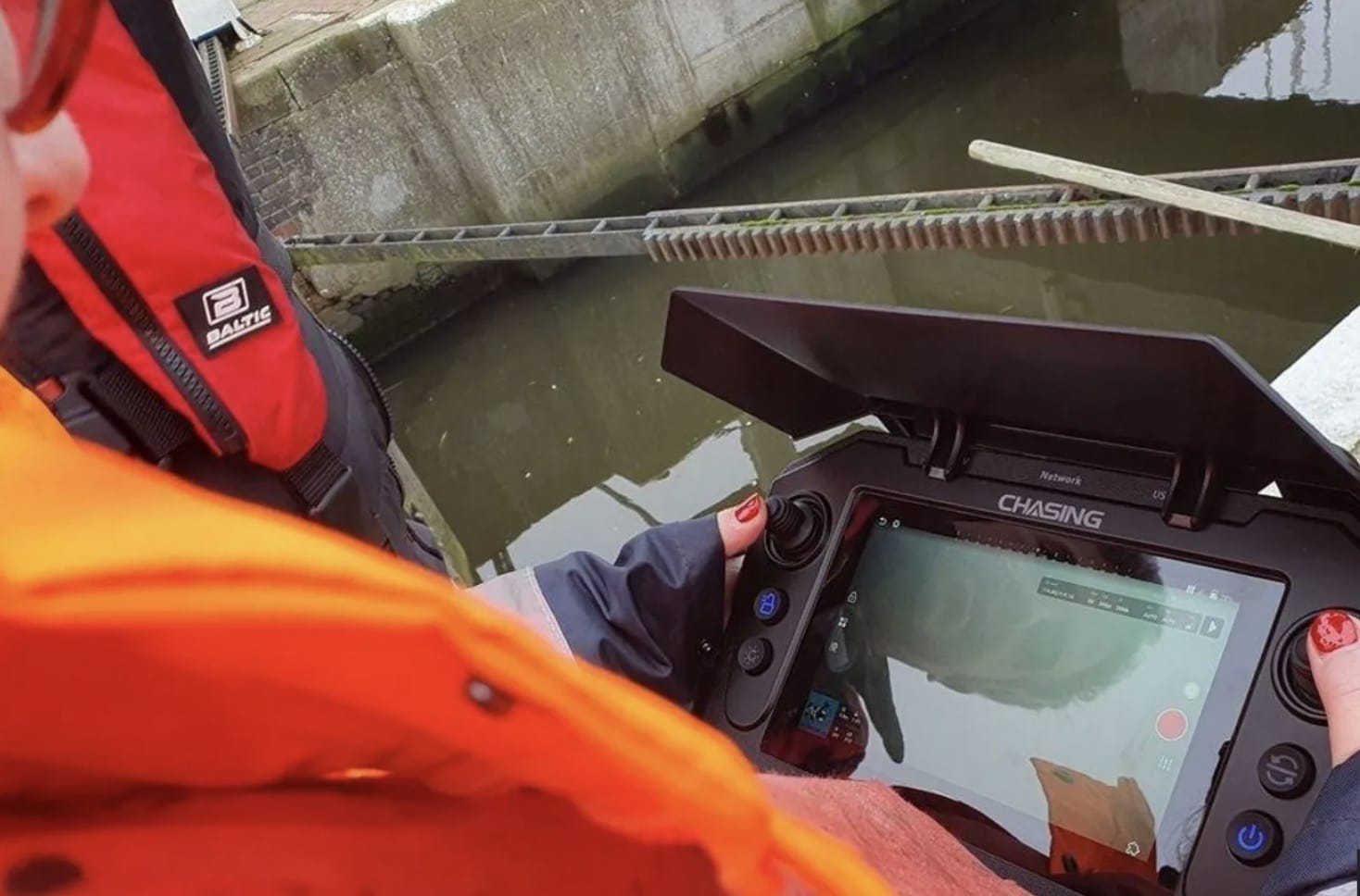 Operator in an orange flotation suit holds a Chasing ROV controller beside a waterway.