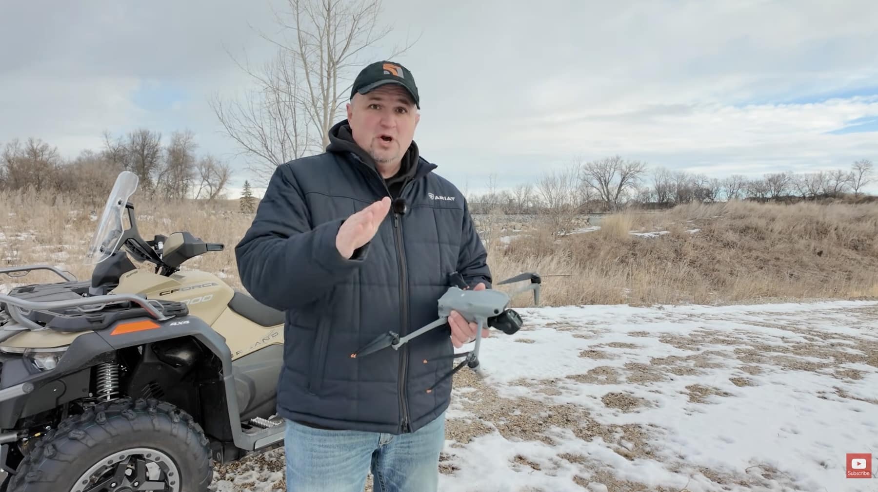 Presenter holds a folded DJI drone beside an ATV in a snowy field.