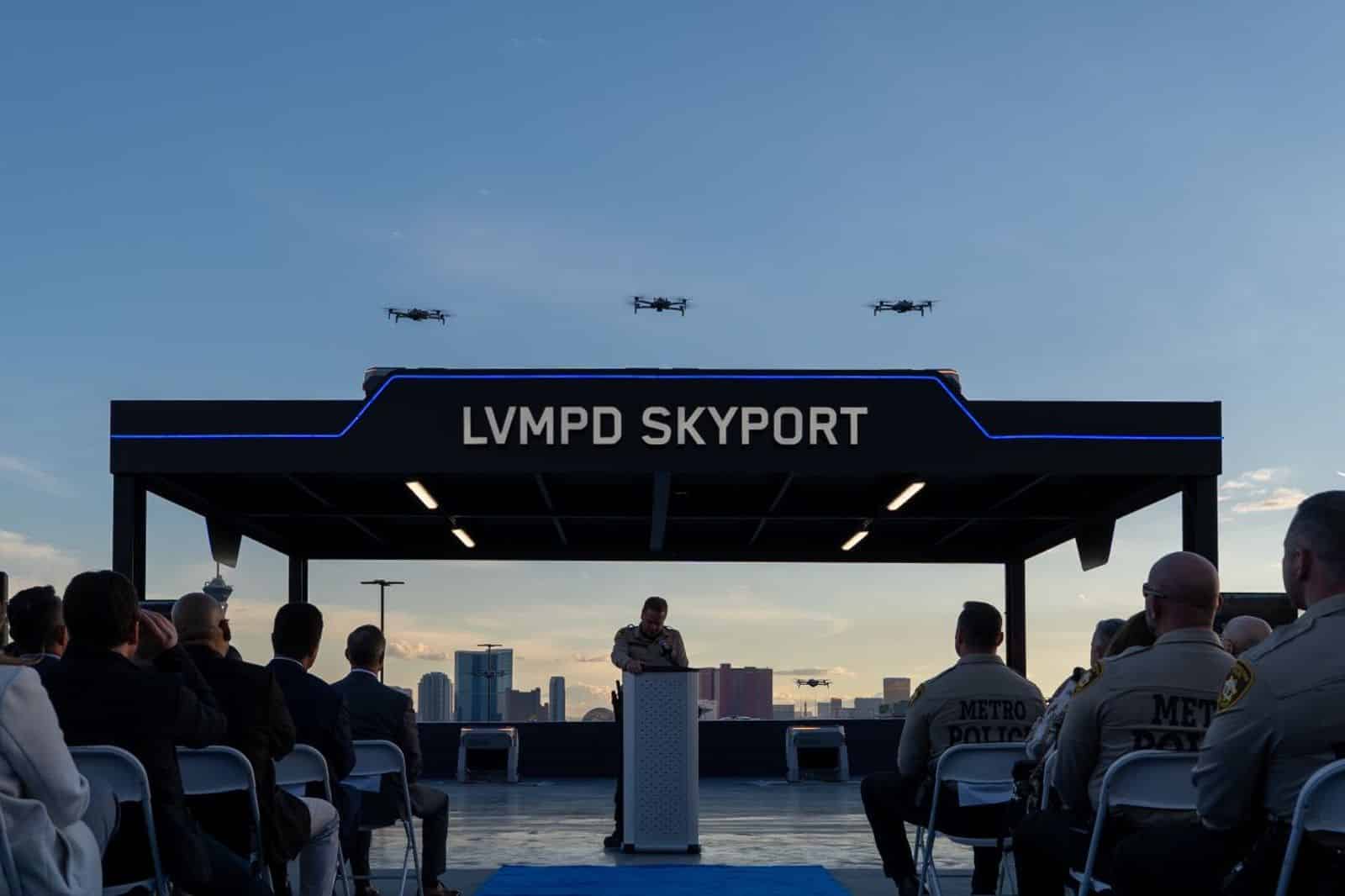 Three drones hover above the LVMPD Skyport as an officer addresses a seated audience.