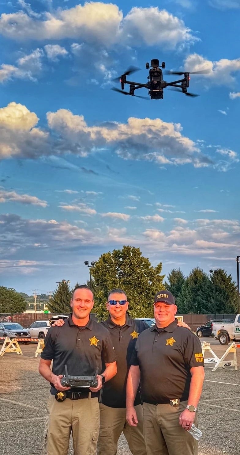 Three sheriff deputies pose with a controller as a small drone hovers above them.