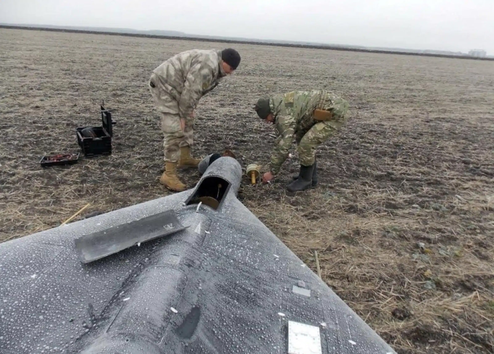 Two soldiers in camouflage prepare a grey delta-wing strike drone on frozen farmland.