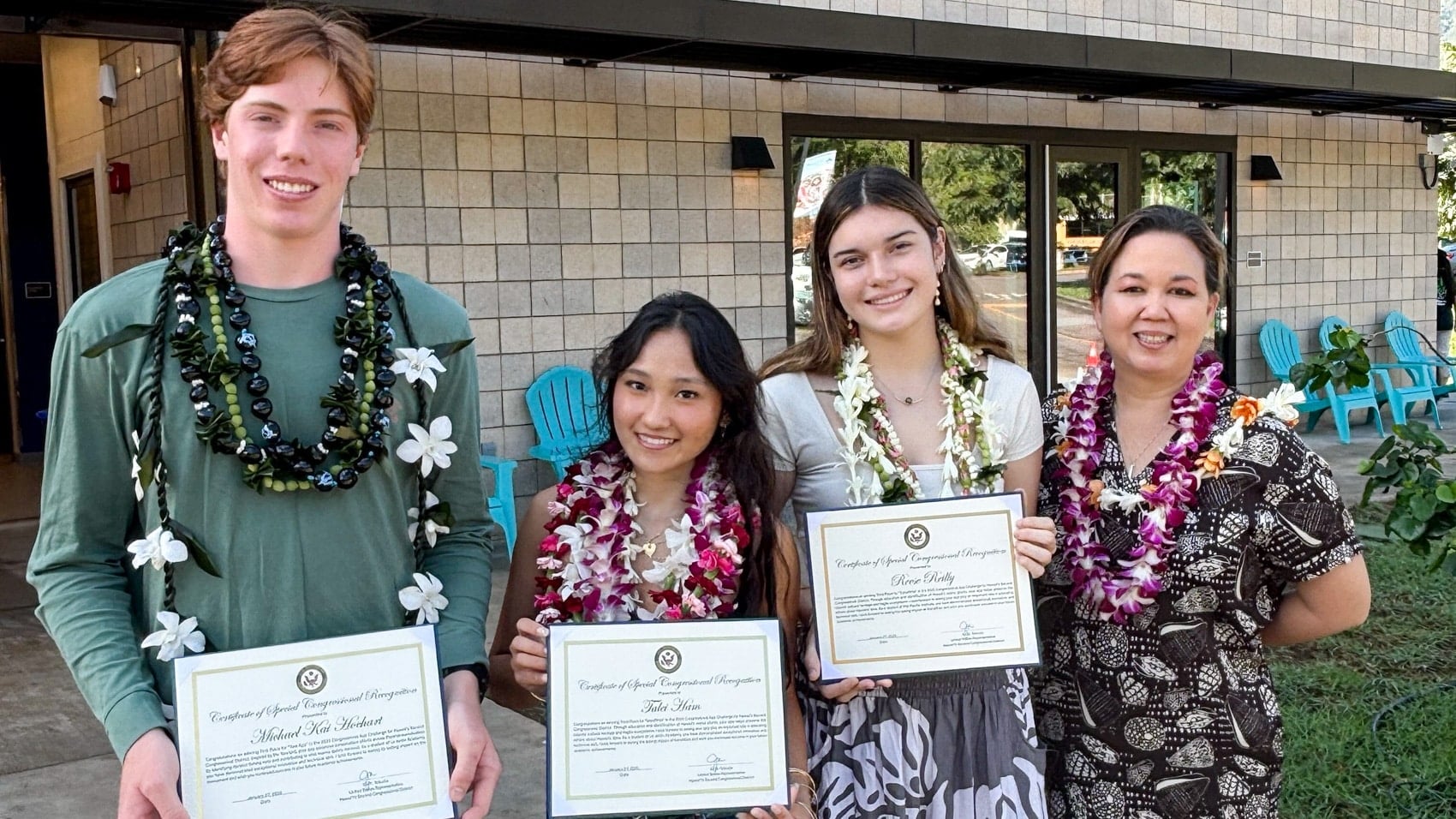 App Challenge winners hold certificates while wearing flower leis outside a school.