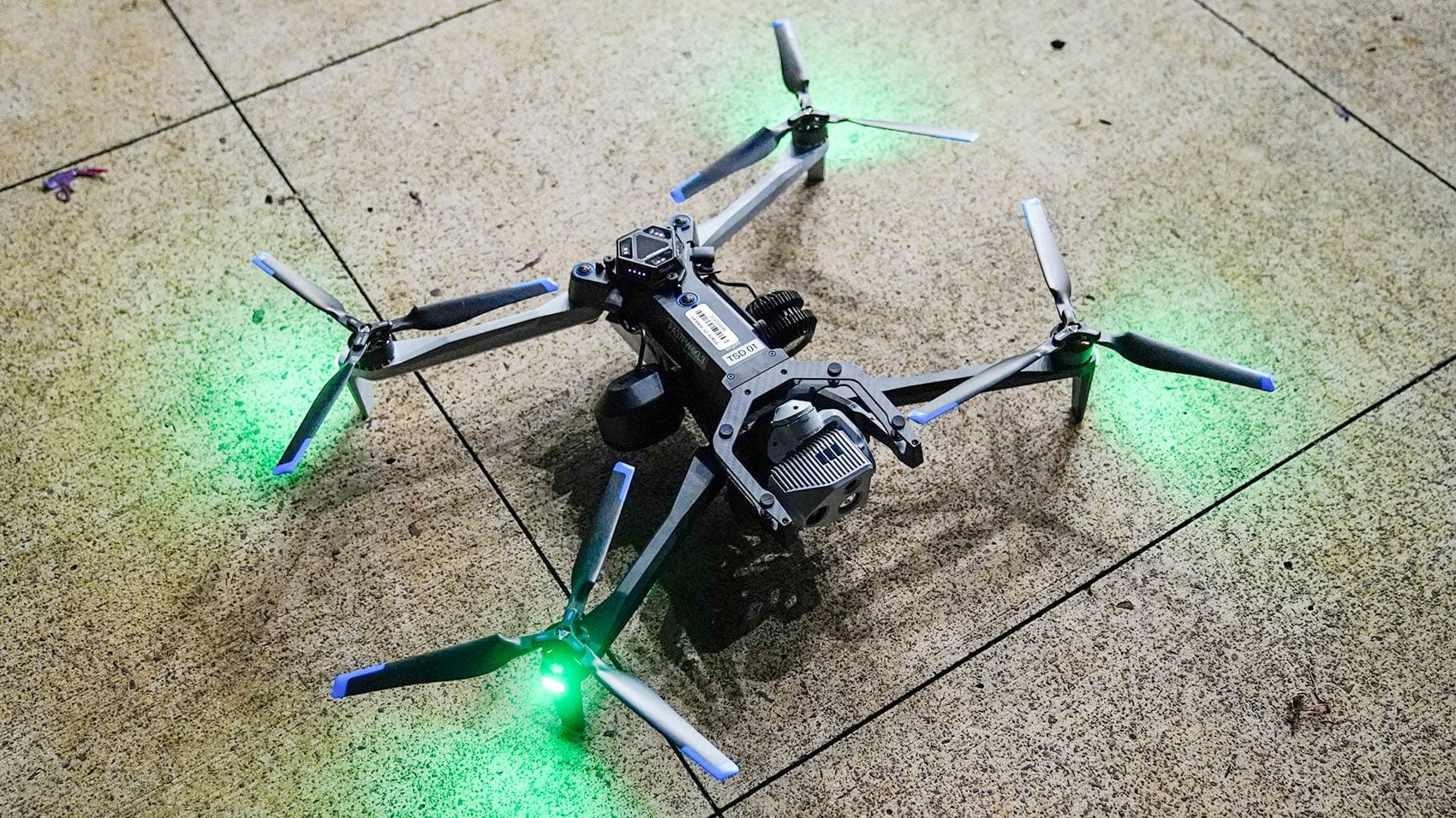 Folding quadcopter with glowing green navigation lights rests on tiled pavement at night.
