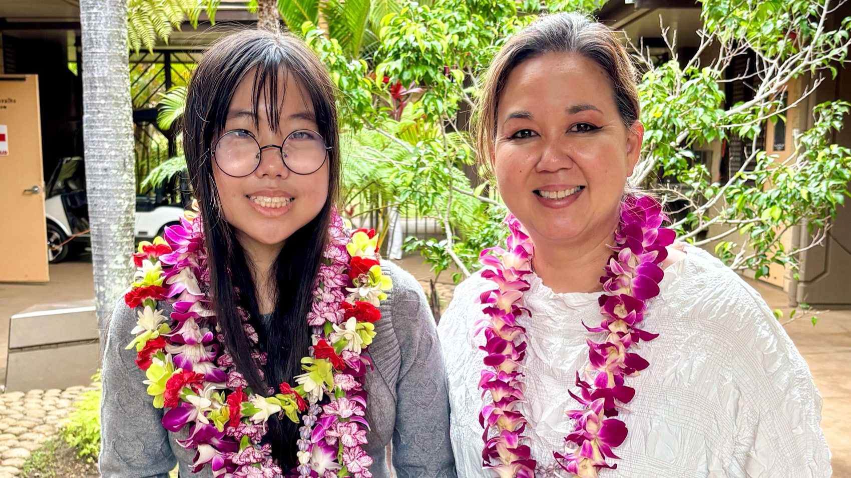 Two award recipients wearing flower leis pose together in dappled sunlight.