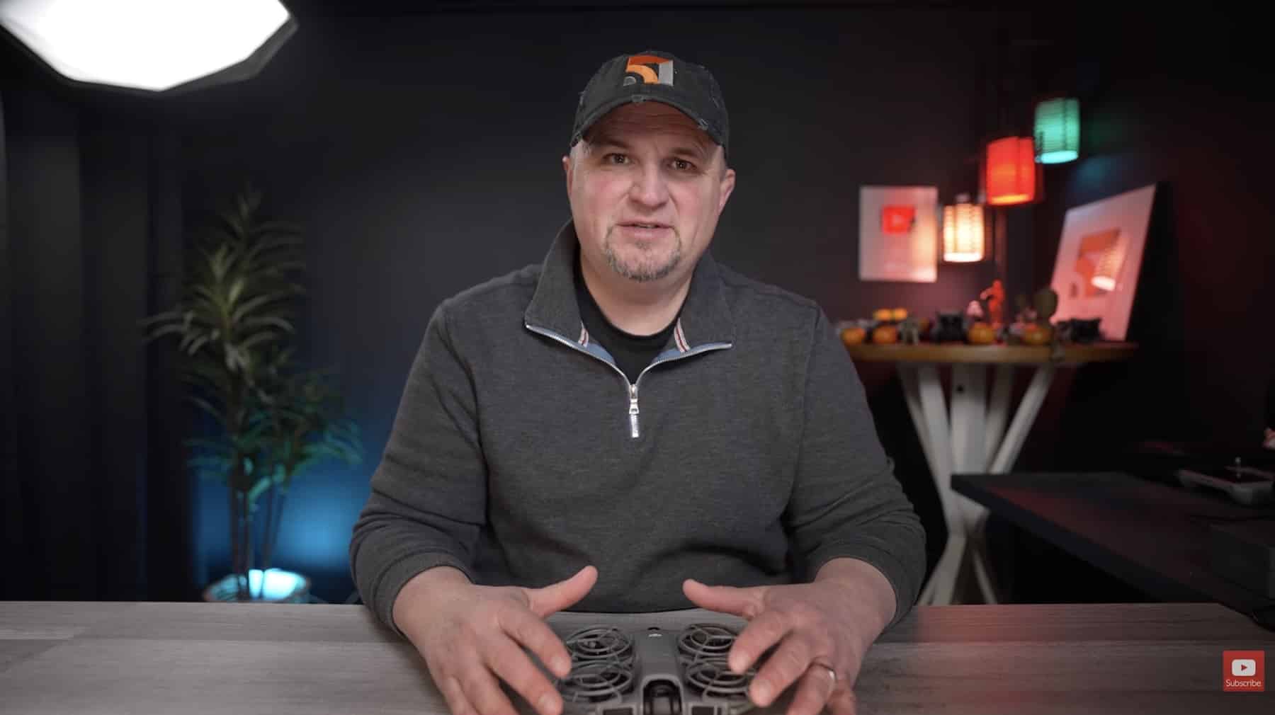 Video host seated at a desk holding a drone controller while talking to camera.