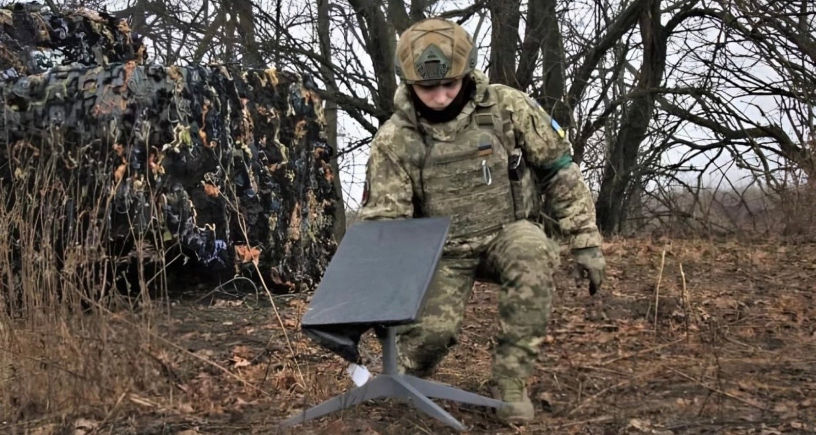 A Ukrainian soldier sets up a Starlink dish on frozen ground beside a camouflaged position.