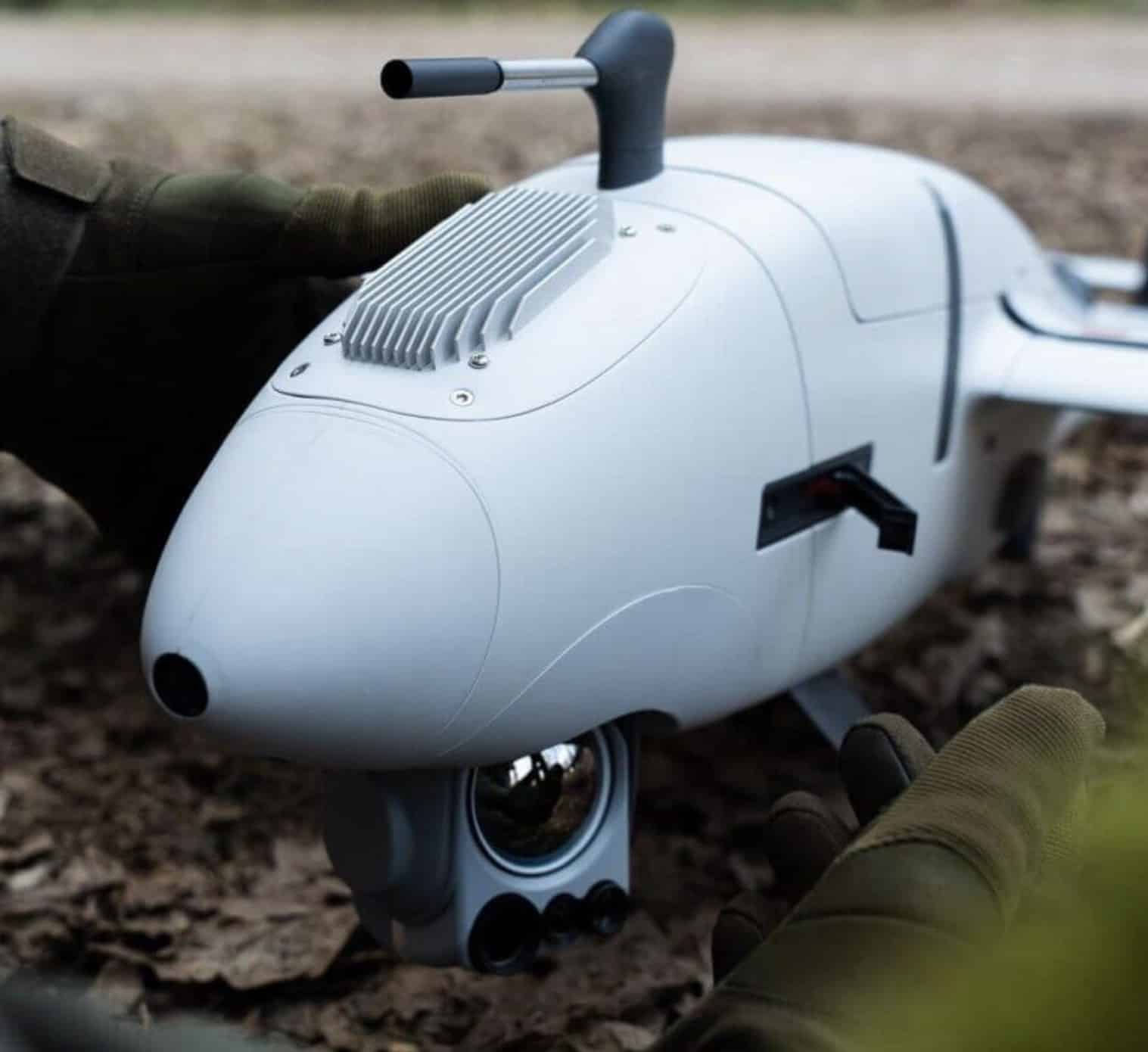 Gloved hands hold a white reconnaissance drone fuselage with its sensor turret.