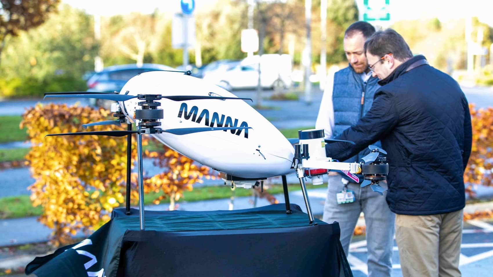 White Manna delivery drone on a stand as two men inspect it outdoors.