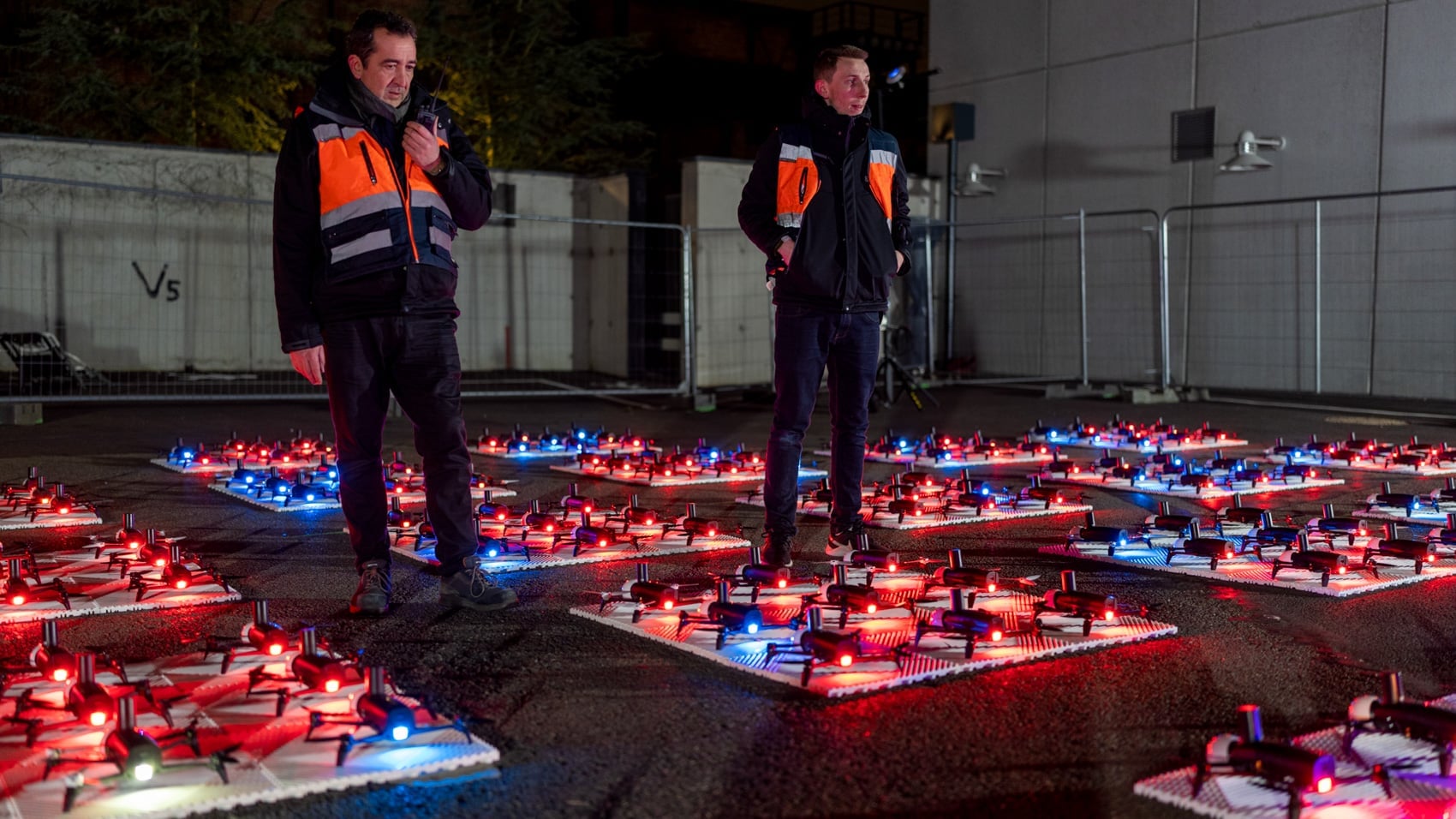 Crew in hi-vis vests stand among grids of glowing show drones before a night launch.