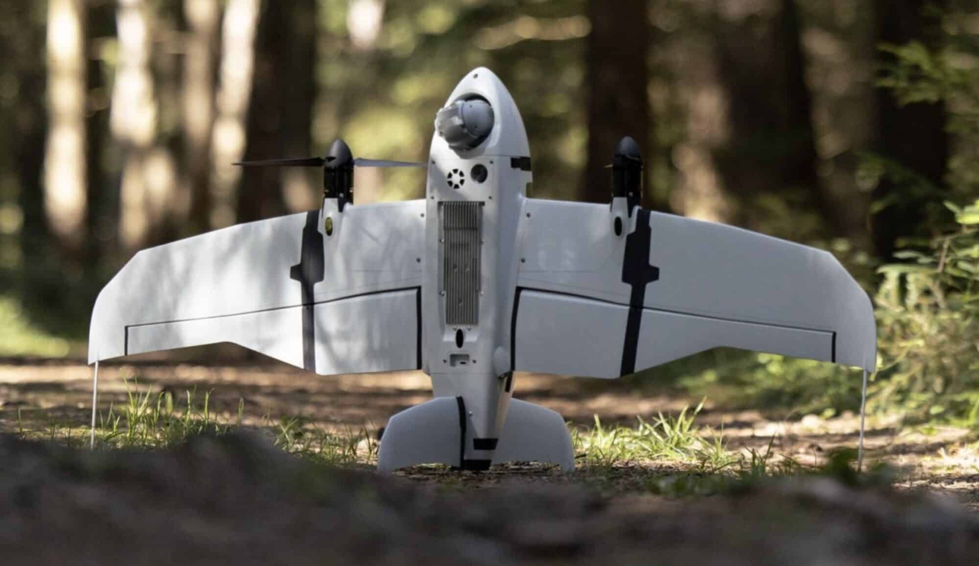 White fixed-wing reconnaissance drone resting on forest ground between pine trunks.