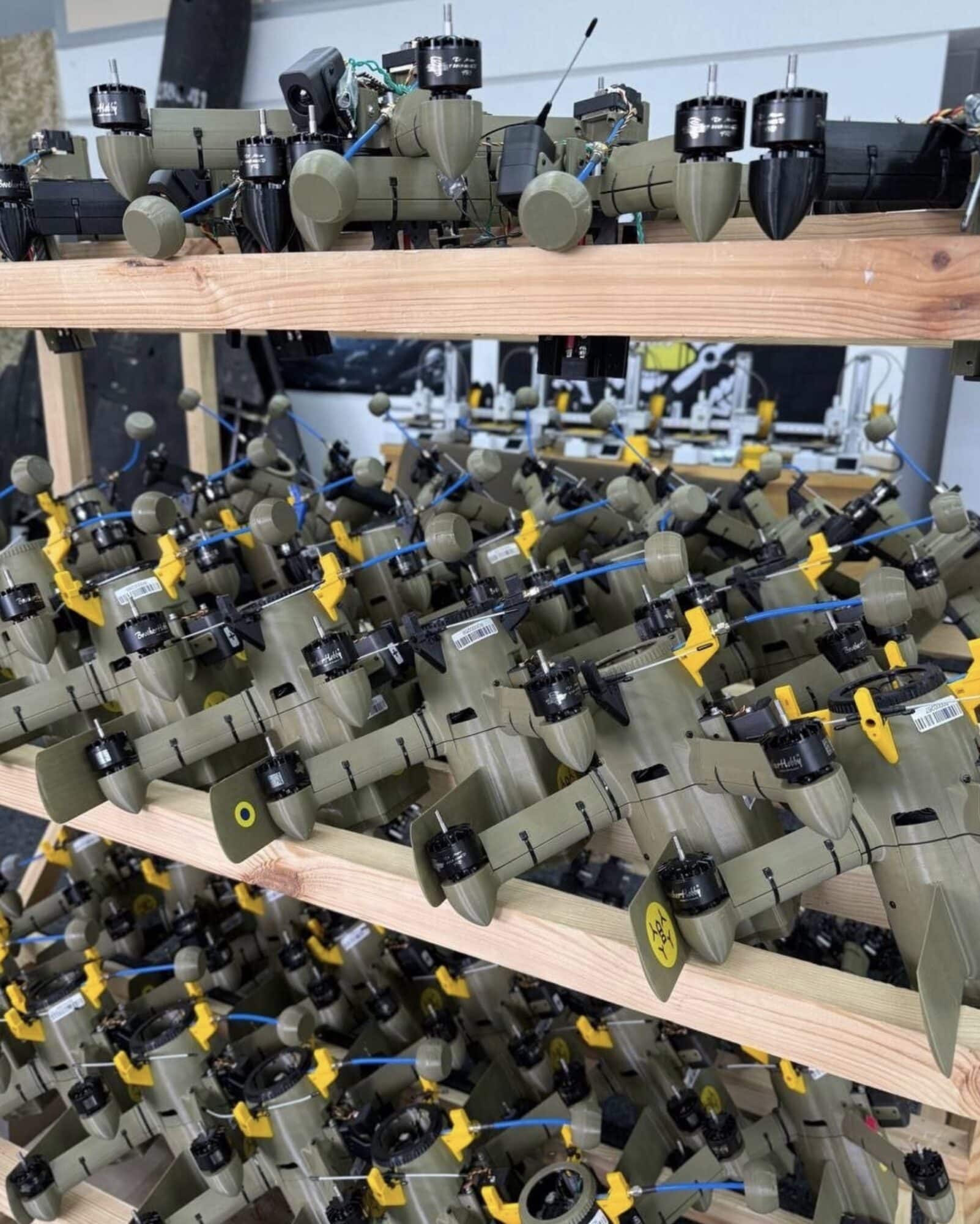 Rows of military drones with yellow fins stacked on wooden workshop shelves.
