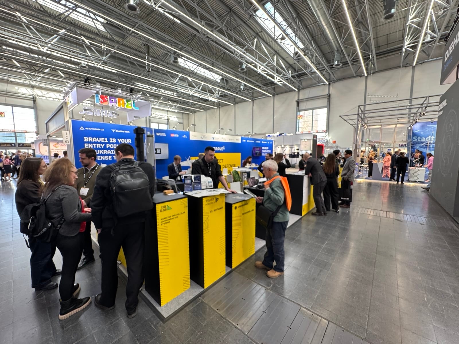 The Brave1 booth banner inviting visitors to Ukraine's defense tech ecosystem, with yellow kiosks.