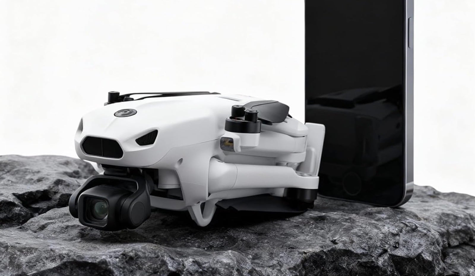 Folded white camera drone propped against a smartphone on a rock for scale.