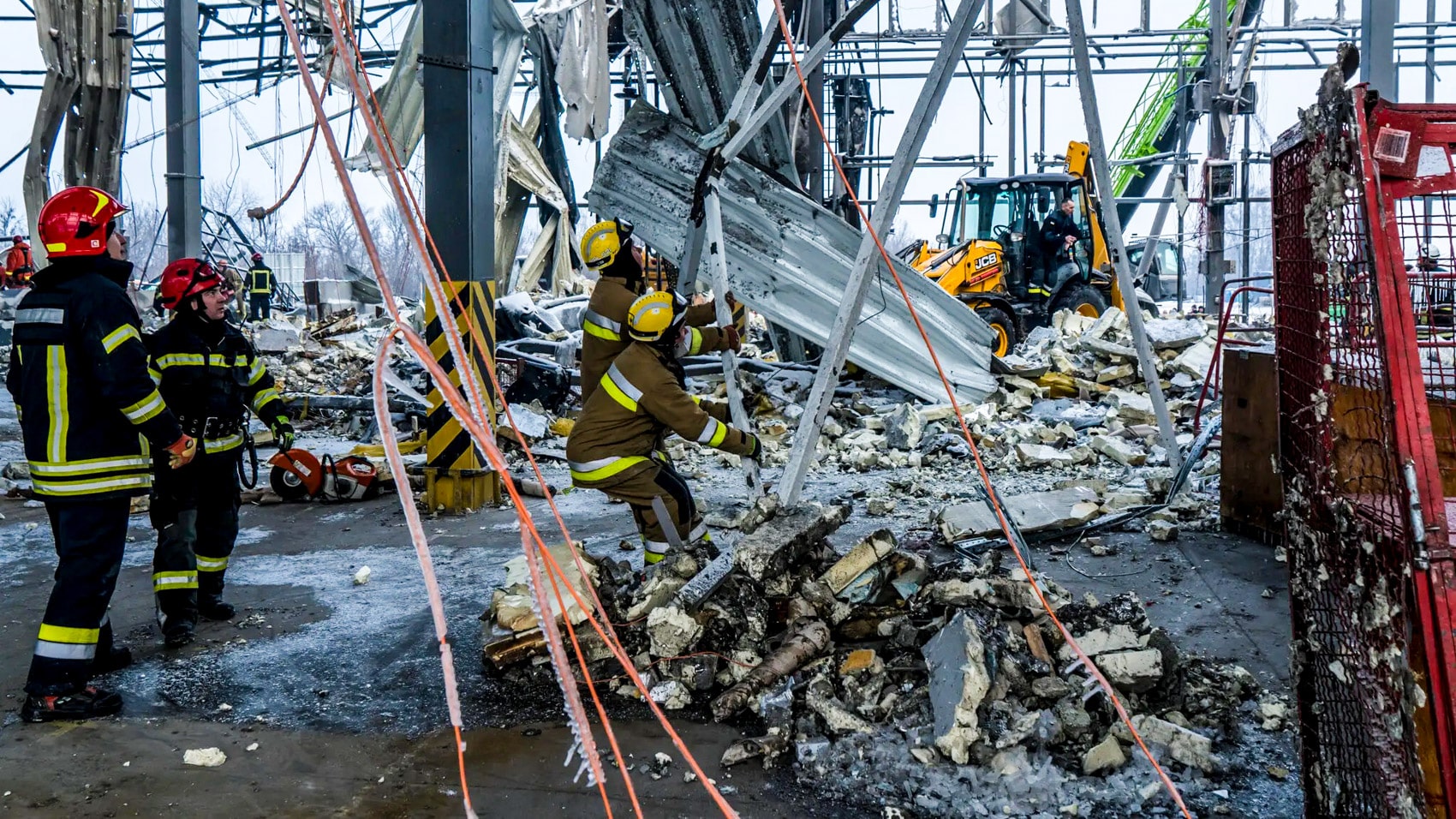 Firefighters climbing through rubble at a collapsed steel and concrete logistics terminal.
