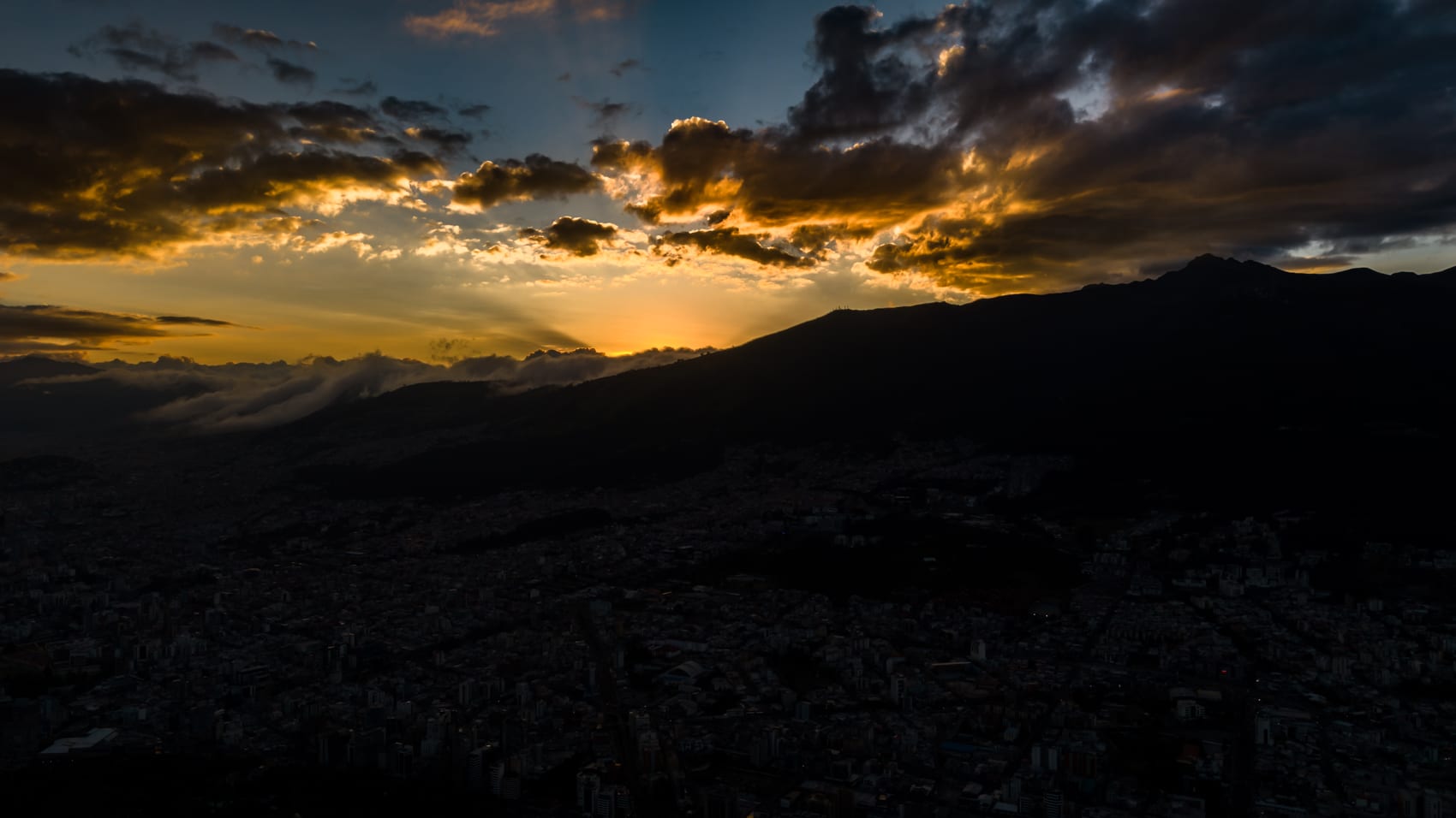 Aerial sunset over a dark mountain ridge with heavy clouds and golden light.