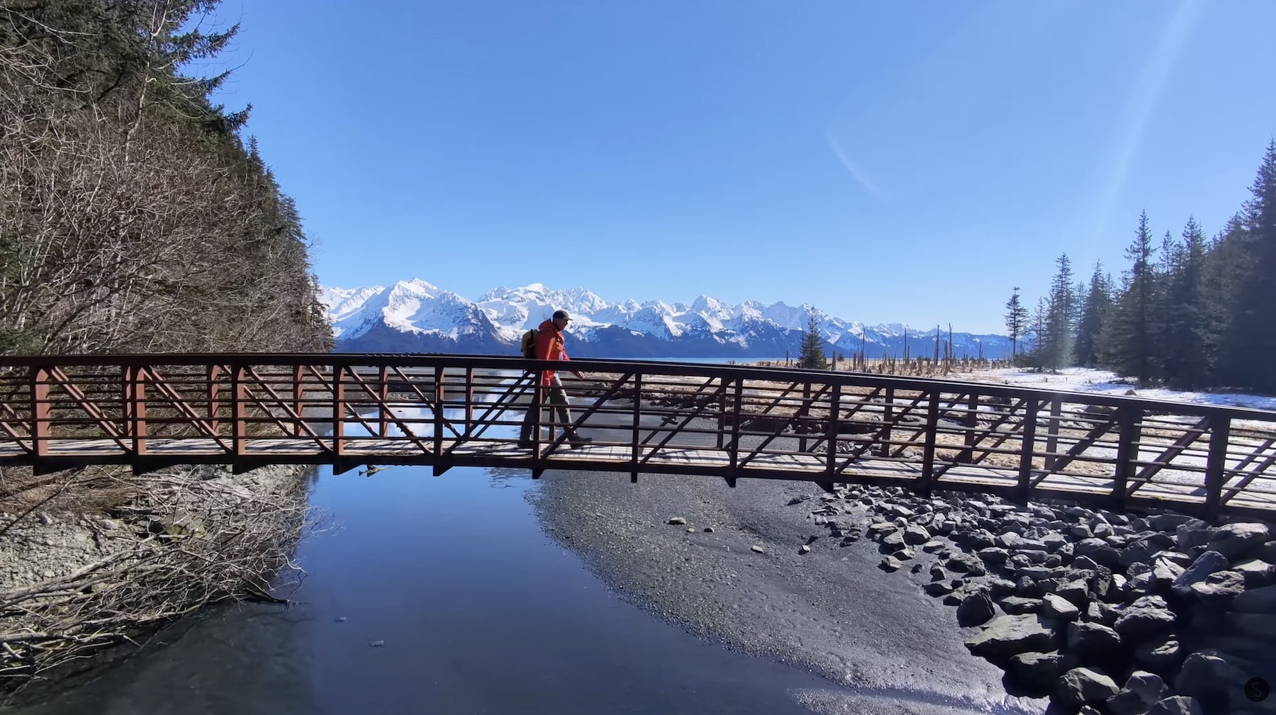 A hiker crosses a wooden footbridge over a creek below snowy Alaskan peaks.