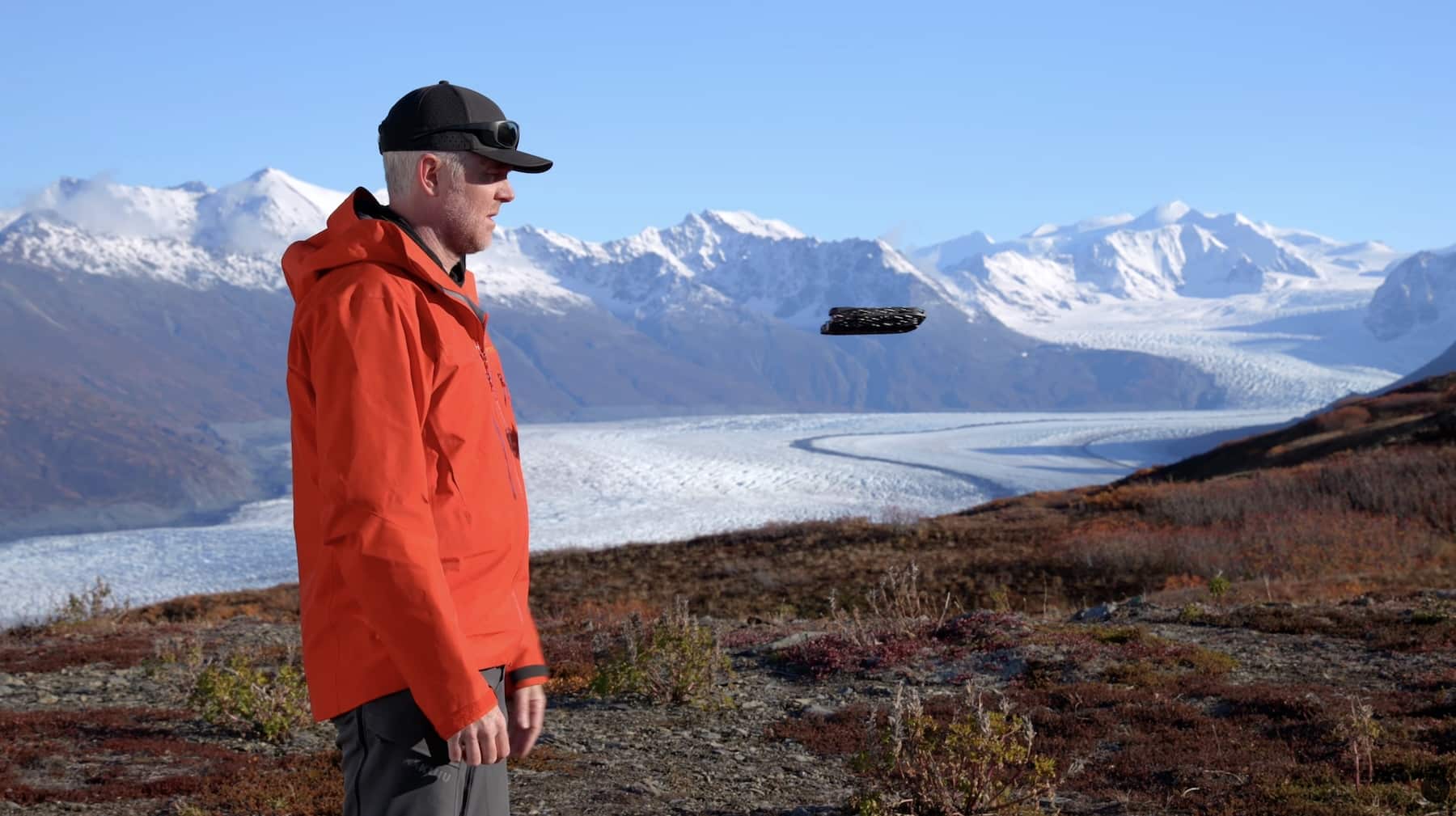 A hiker in an orange jacket watches a small drone above an Alaskan glacier.