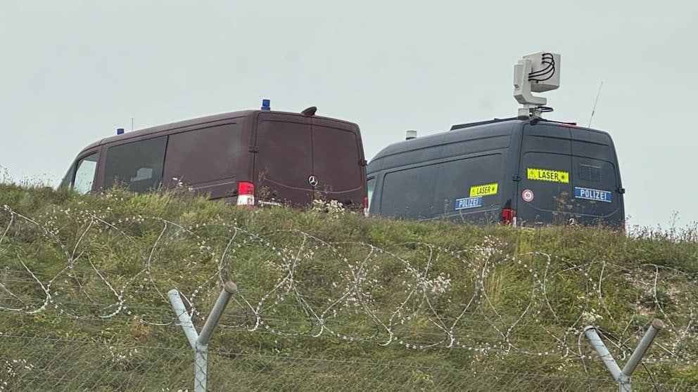 Two police vans behind razor wire, one carrying a mast-mounted laser detection head.