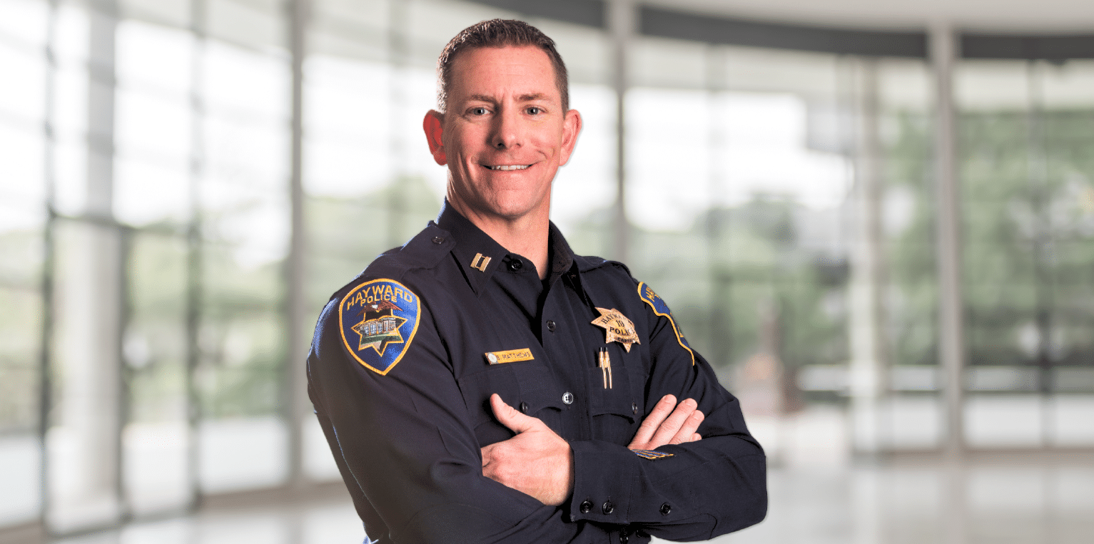 Uniformed Hayward police officer standing with arms crossed in a bright lobby.