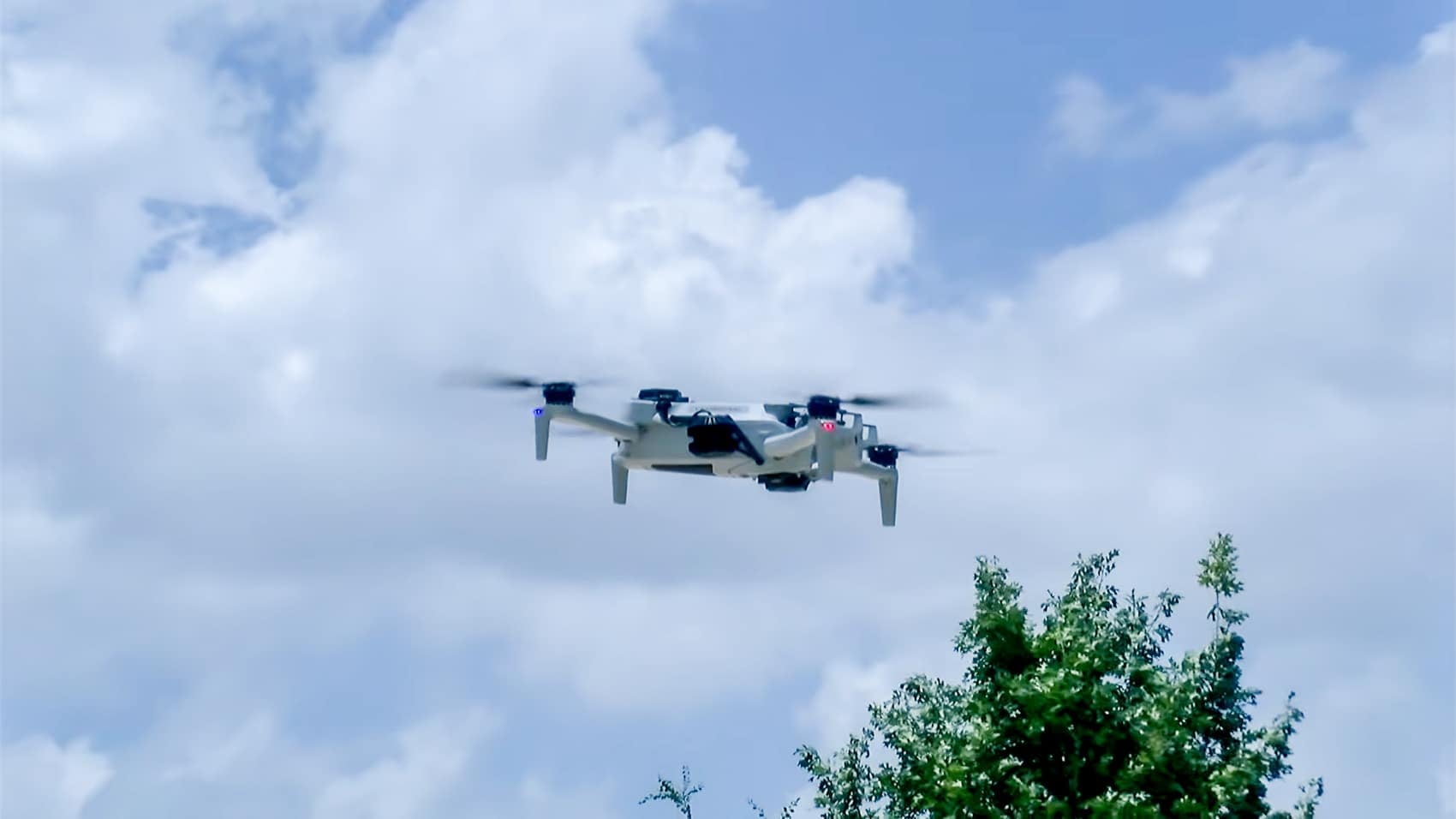 Small folding quadcopter climbing against clouds above a leafy treetop.