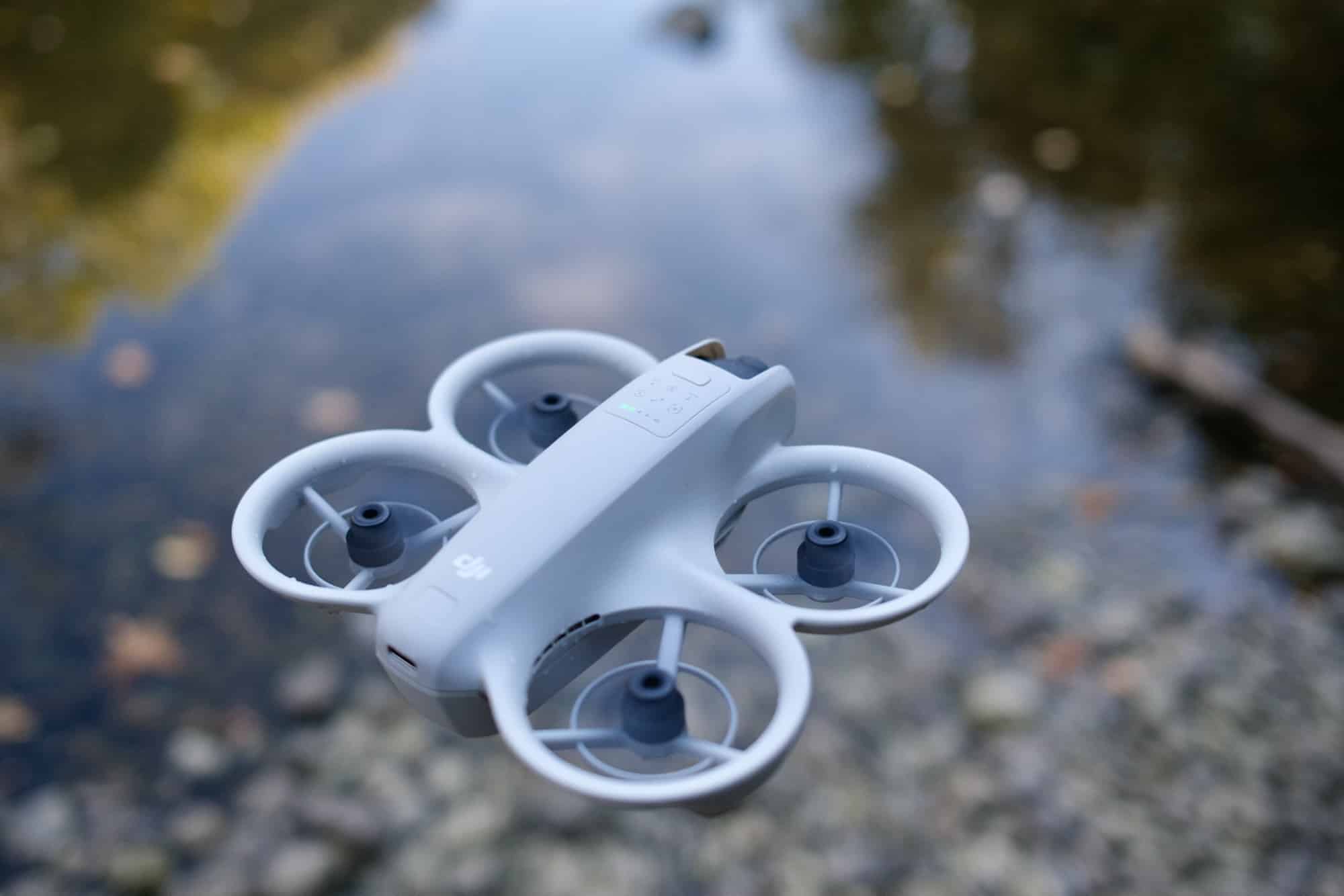 DJI Neo hovering low above a shallow stream lined with smooth pebbles.