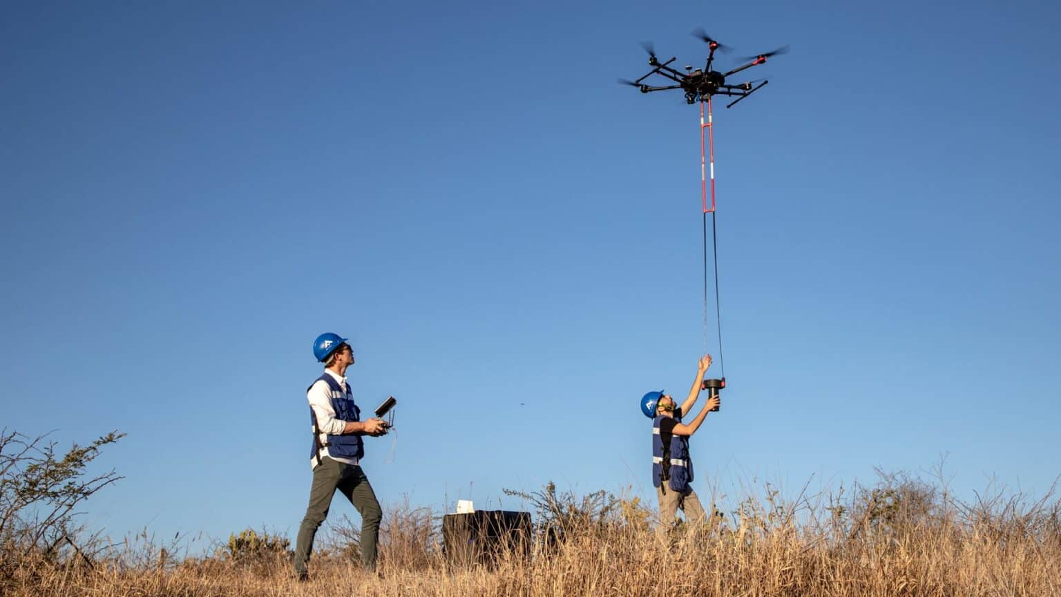 Two crew members guide a hexacopter towing a suspended magnetometer over scrubland.
