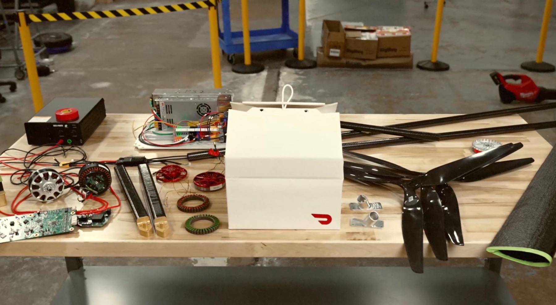 Drone motors, propellers, carbon fiber frame tubes, electronics and a DoorDash branded delivery bag arranged on a workbench