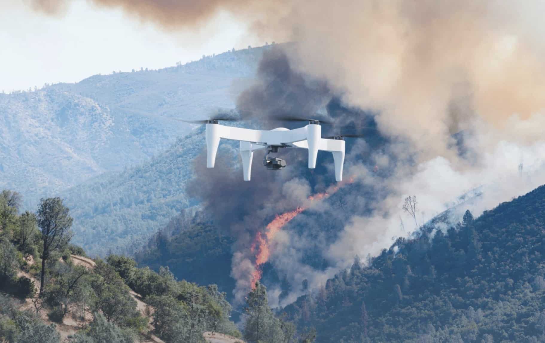 White quadcopter flying above a smoking hillside wildfire in the mountains.