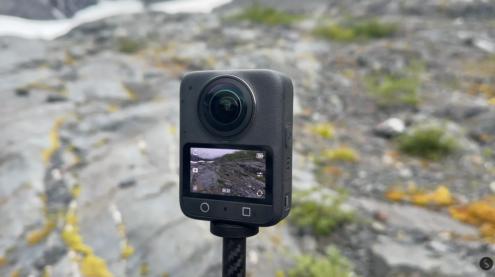 Black 360-degree action camera mounted on a carbon fiber selfie stick, its rear screen showing an 8K30 preview of the rocky alpine landscape behind it