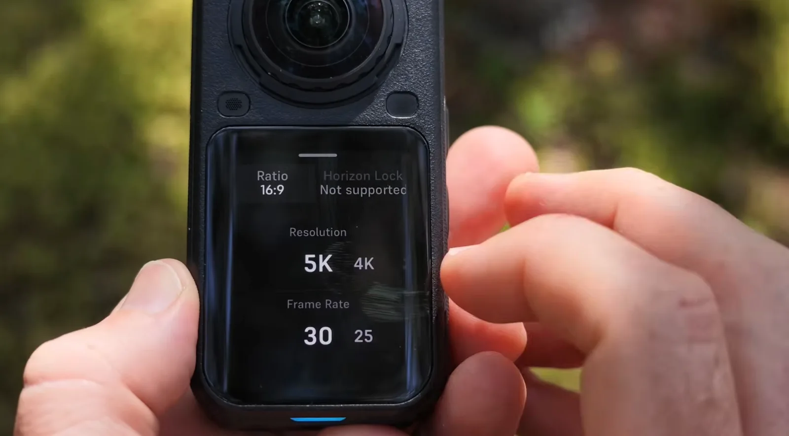 Hands holding a black 360-degree action camera, its touchscreen showing the video settings menu with 16:9 ratio, 5K resolution and 30 fps selected