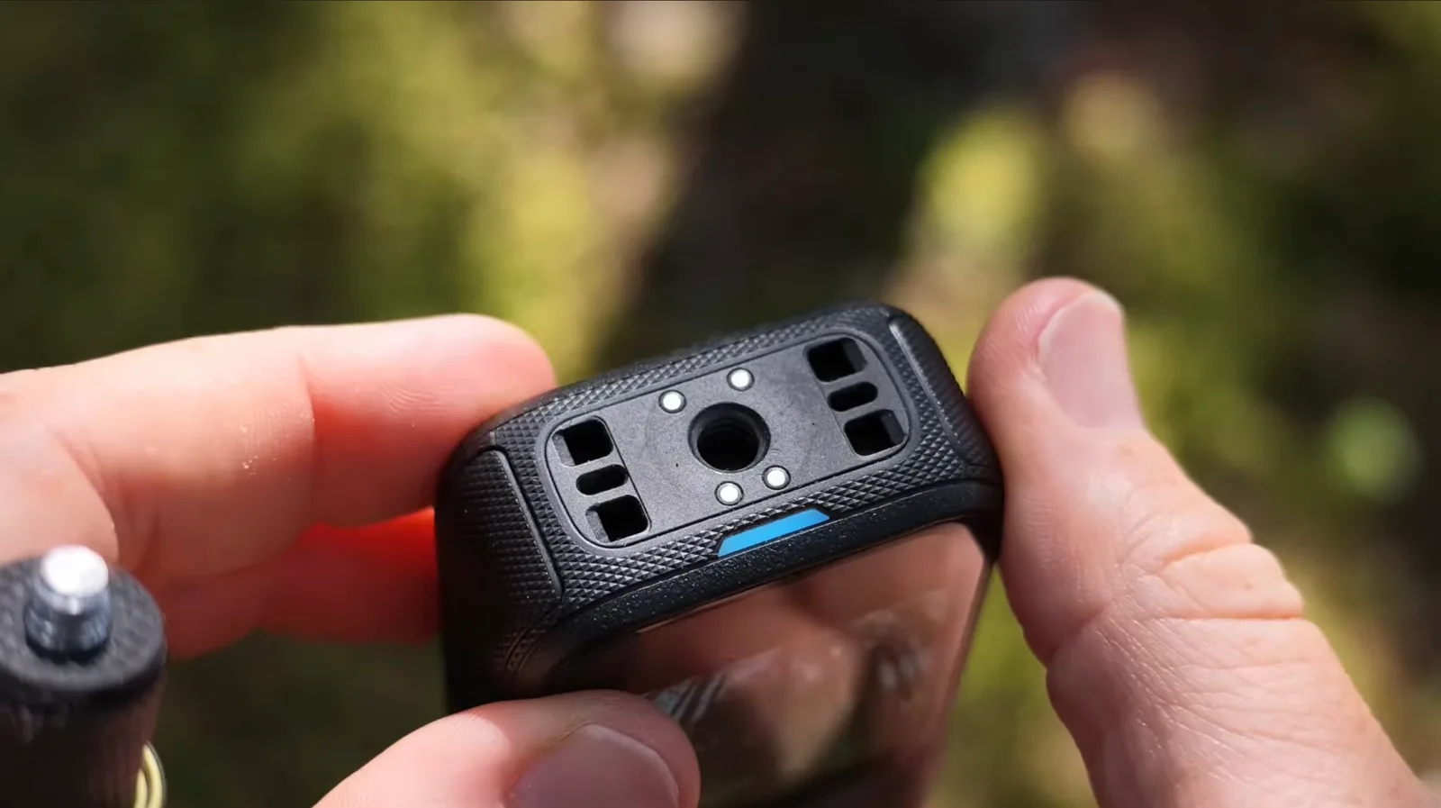 Close-up of two hands holding a rugged black action camera, showing its textured underside with a threaded tripod socket, magnetic contact pads and a blue indicator strip.