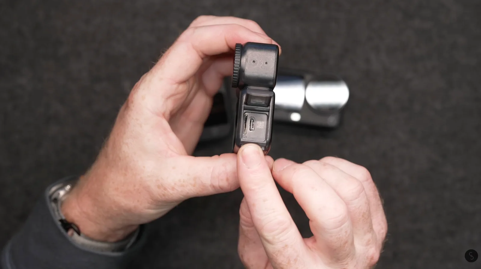 Hands holding a small black action camera with its side port door flipped open, revealing the USB-C charging port and microSD card slot