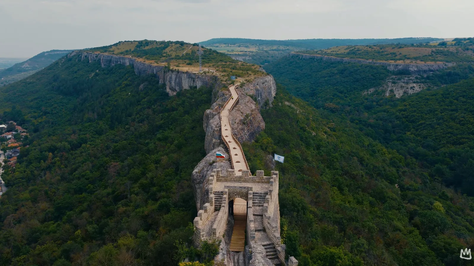 Aerial view of a medieval hilltop fortress gate with a long wooden walkway winding along a forested limestone ridge