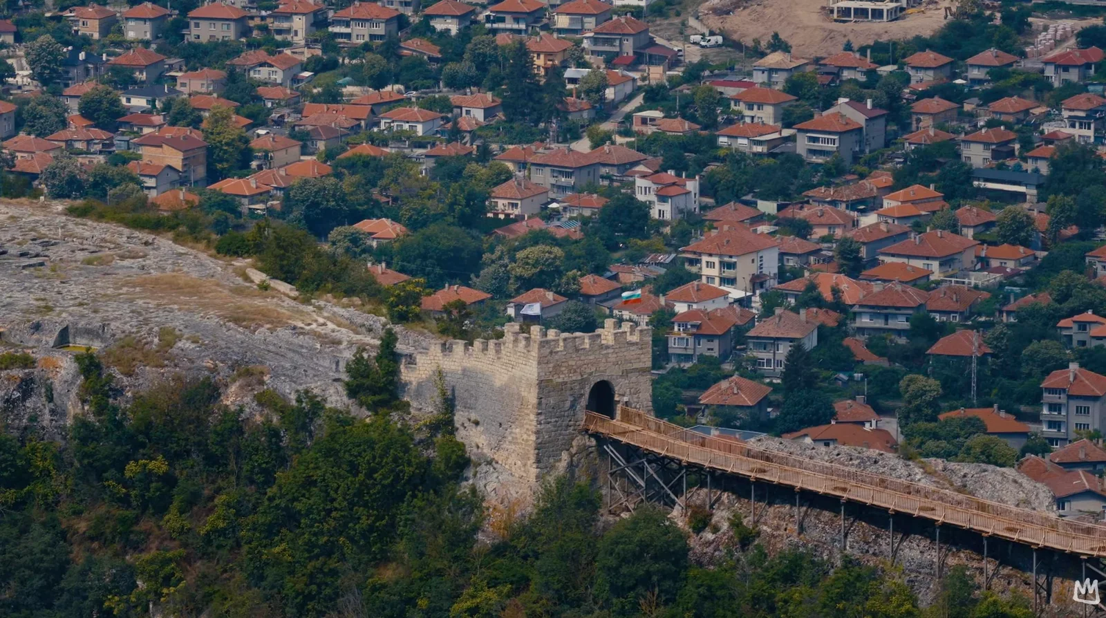 Aerial view of a stone medieval fortress gate flying the Bulgarian flag, connected by a long wooden bridge to a clifftop, with a red-roofed town spread across the hillside behind it