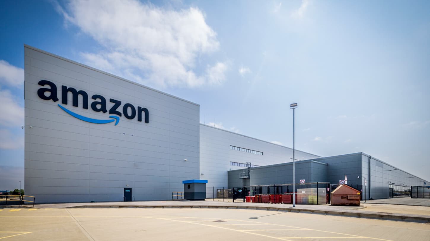 The exterior of Amazon's fulfillment center in Darlington, County Durham, a long grey warehouse with the Amazon logo on its end wall under a blue sky