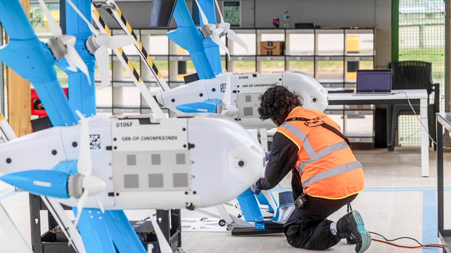 Two Amazon Prime Air MK30 drones parked side by side inside the Darlington hangar, each marked with a UK operator ID, as a technician in a Prime Air hi-vis vest kneels beside one