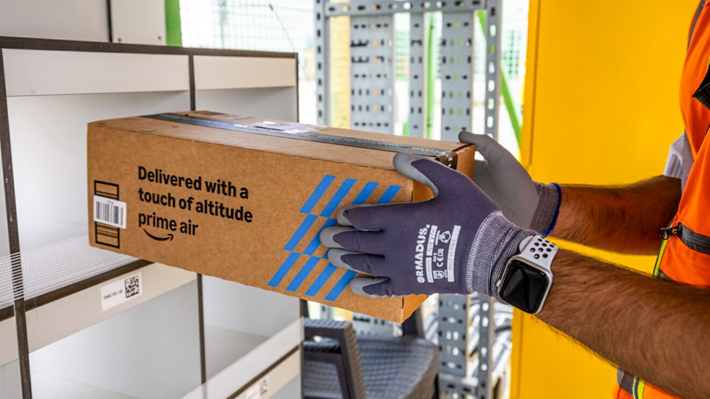 A gloved Prime Air worker slides a long cardboard Amazon box printed with the words delivered with a touch of altitude into a sorting shelf