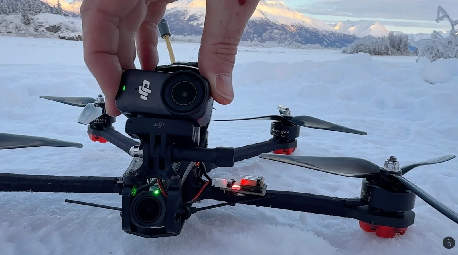 Hand mounting a DJI action camera on top of a custom long-range FPV quadcopter resting in snow, with sunlit mountains behind