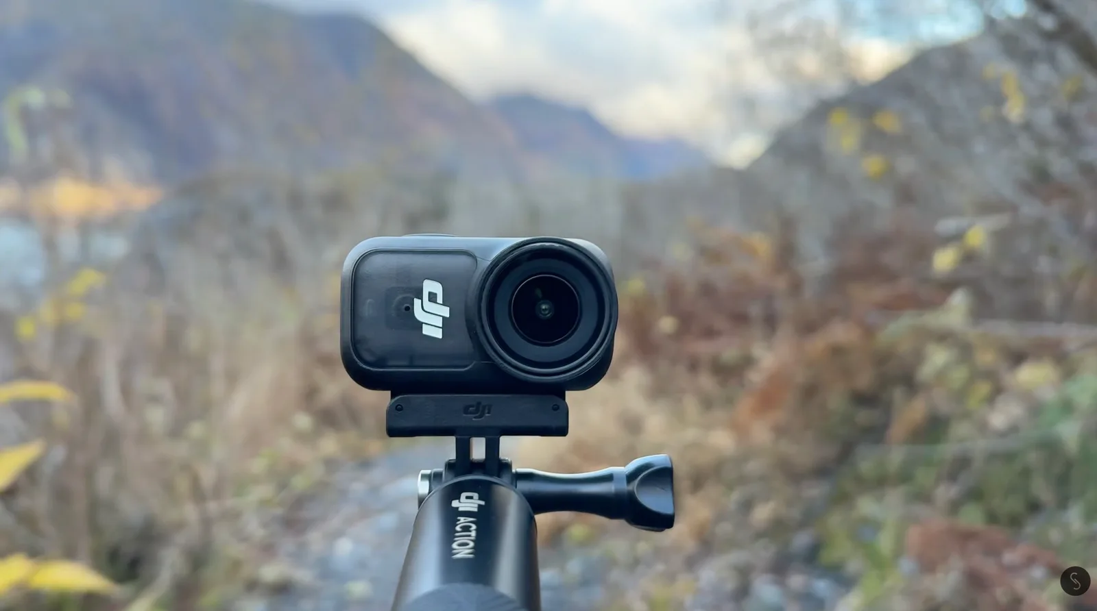 Compact DJI action camera mounted on a selfie stick handle in front of a blurred autumn mountain valley